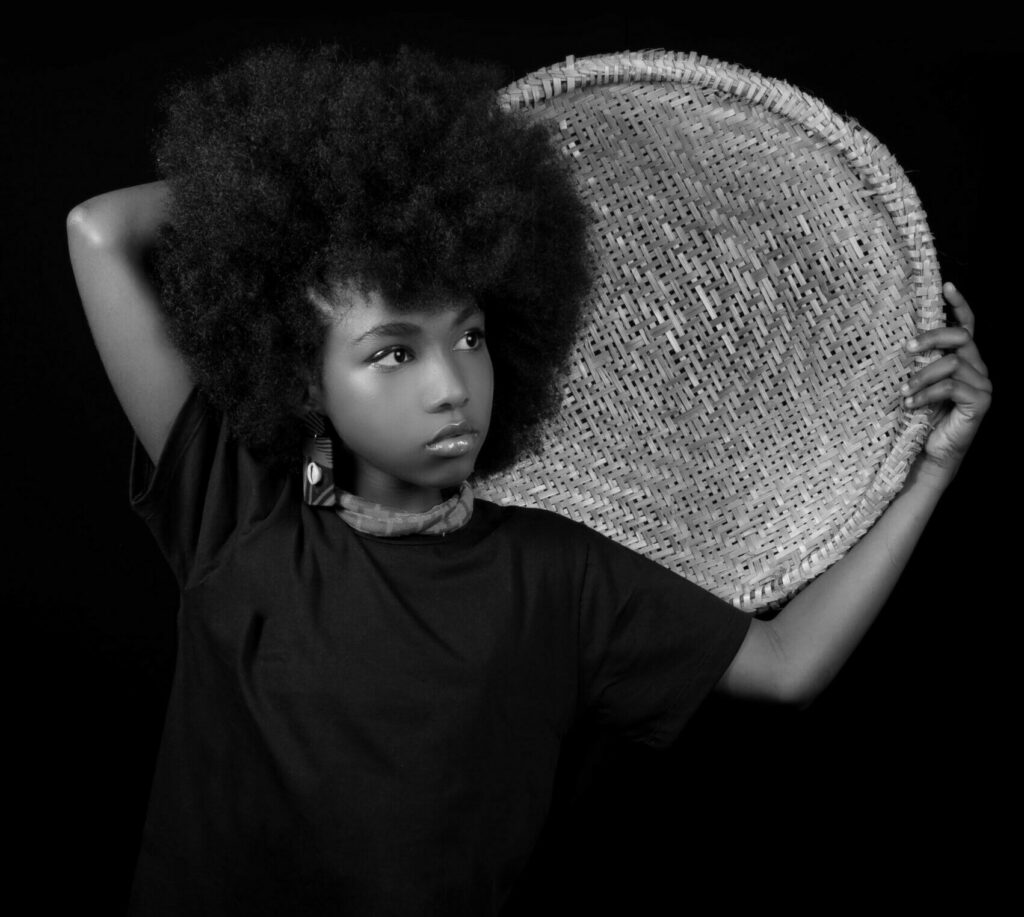 Monochrome portrait of a young girl with afro hairstyle holding a woven tray against a black background.